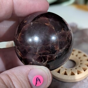 May include: A polished, dark burgundy sphere with a cracked, textured surface. The gemstone is held in a hand, with a pink sticker labeled "A" visible. A carved wooden disc is in the background.