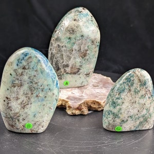 May include: Three smooth, polished, green and white, freeform, natural stones. The stones are arranged in a row on a brown surface.