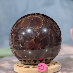 May include: A polished, dark brown gemstone sphere with a textured surface. The sphere is resting on a wooden stand. The gemstone has a reflective quality, with lighter brown and gold inclusions.