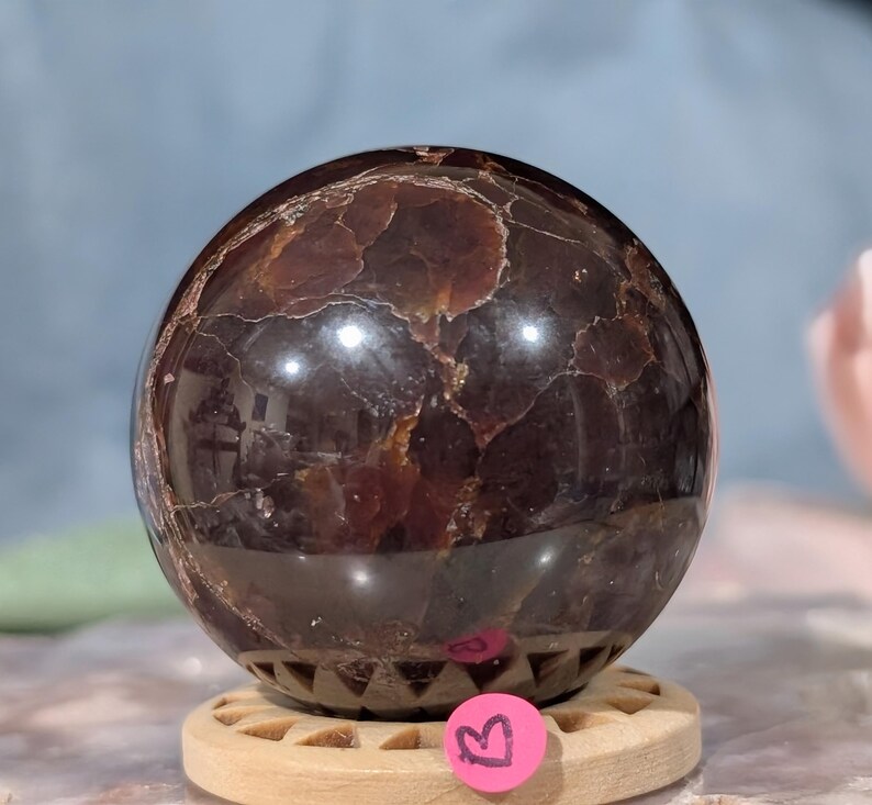 May include: A polished, dark brown gemstone sphere with a textured surface. The sphere is resting on a wooden stand. The gemstone has a reflective quality, with lighter brown and gold inclusions.