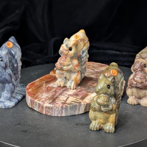 May include: Four carved stone squirrel figurines in various colors, including blue, tan, and brown. Each squirrel is holding a nut. The figurines are displayed on a black surface and a wooden slab.