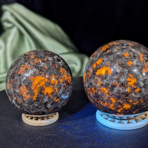 May include: Two polished spheres of black and orange stone, each sitting on a white stand. The spheres have a speckled pattern of orange and black.