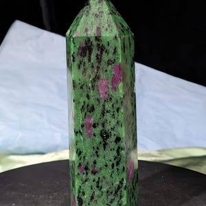 May include: A green and pink ruby zoisite crystal obelisk. The polished stone has a rectangular shape with a pointed top. The green base color is speckled with black and pink inclusions.