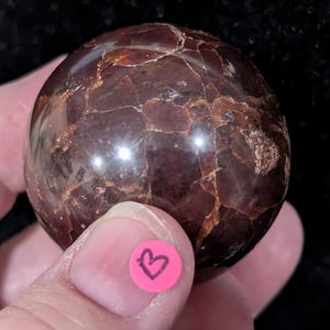 May include: A polished, spherical gemstone with a deep burgundy color and intricate veining. The surface has a glossy sheen, and the stone is held in a hand. A small pink sticker with a black heart is visible.