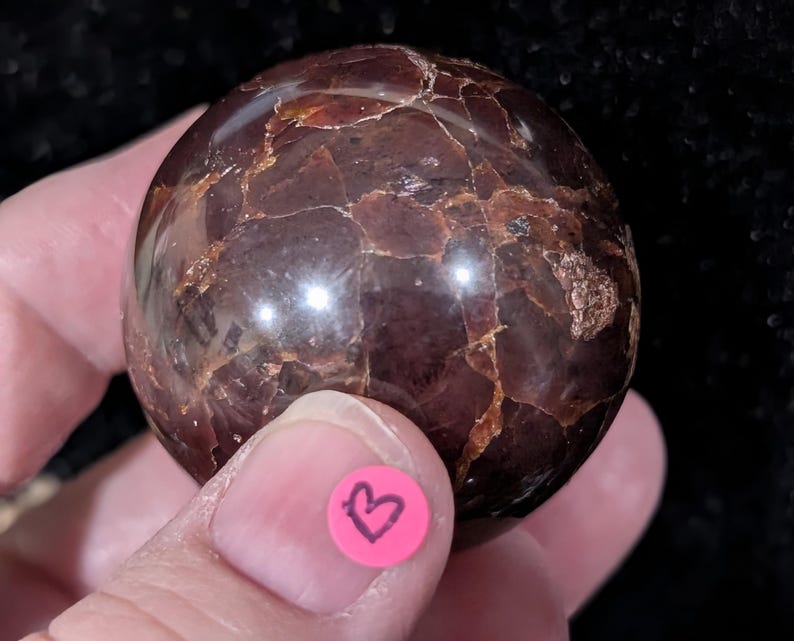 May include: A polished, spherical gemstone with a deep burgundy color and intricate veining. The surface has a glossy sheen, and the stone is held in a hand. A small pink sticker with a black heart is visible.