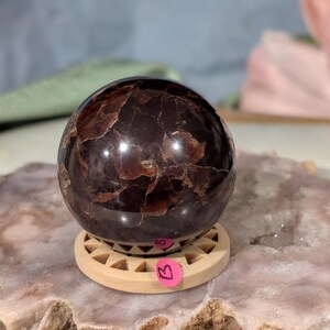 May include: A polished, dark brown gemstone sphere with a textured surface, resting on a small, light-colored wooden stand. The sphere has a reflective quality, and a pink tag is attached to the stand. The sphere is a decorative item.