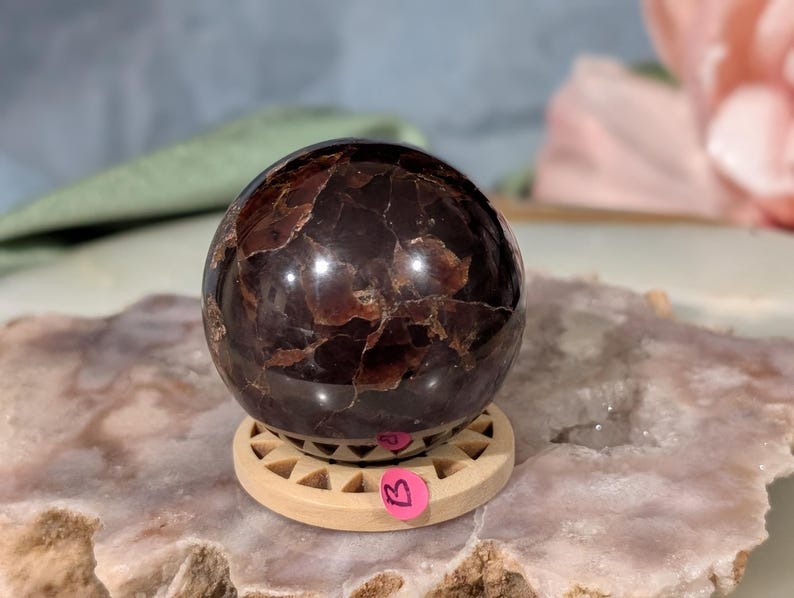 May include: A polished, dark brown gemstone sphere with a textured surface, resting on a small, light-colored wooden stand. The sphere has a reflective quality, and a pink tag is attached to the stand. The sphere is a decorative item.