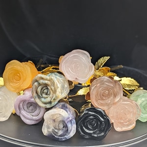 May include: A bouquet of nine rose-shaped crystal carvings in various colors, including pink, green, orange, purple, and black. The roses are arranged on a black surface and have gold-colored stems.