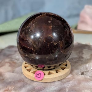 May include: A polished, dark red-brown gemstone sphere, resting on a light wooden stand. The sphere has a smooth, reflective surface with natural inclusions. A small, pink sticker with a letter "B" is affixed to the stand. The sphere is approximately 3 inches in diameter.