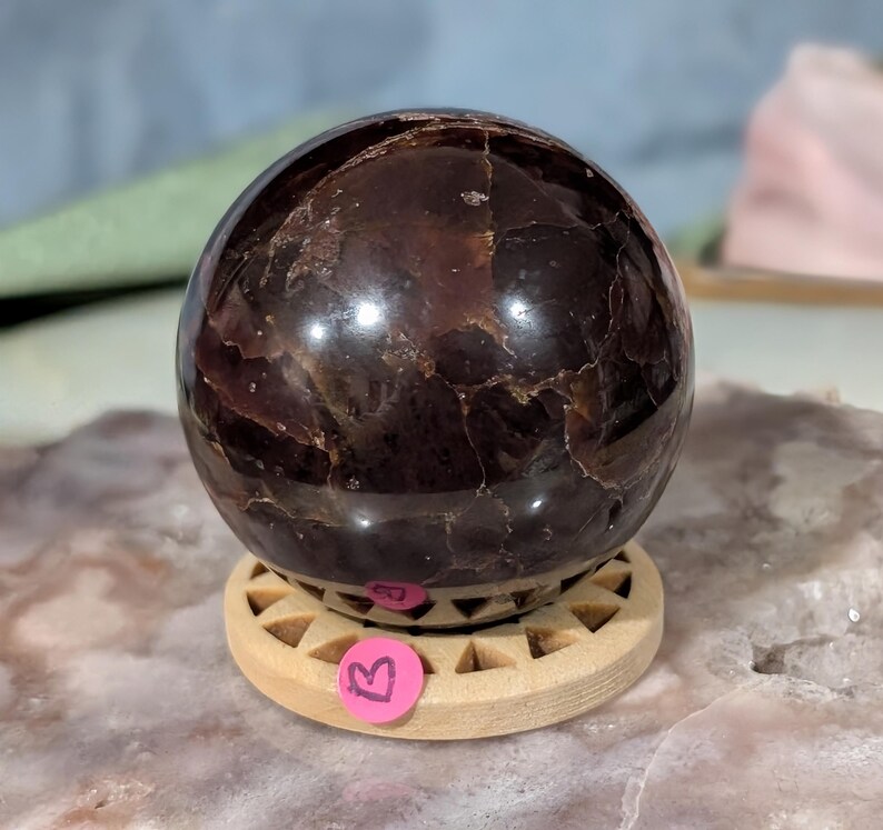 May include: A polished, dark red-brown gemstone sphere, resting on a light wooden stand. The sphere has a smooth, reflective surface with natural inclusions. A small, pink sticker with a letter "B" is affixed to the stand. The sphere is approximately 3 inches in diameter.