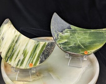 May include: Two crescent-shaped green and white polished stone slabs on clear acrylic stands. The slabs have a natural, striated pattern and are displayed on a white surface.