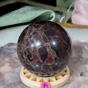 May include: A polished, dark burgundy sphere with a cracked, mosaic-like pattern. The gemstone ball sits on a light wooden stand with triangular cutouts. A small pink sticker with the letter "A" is visible.