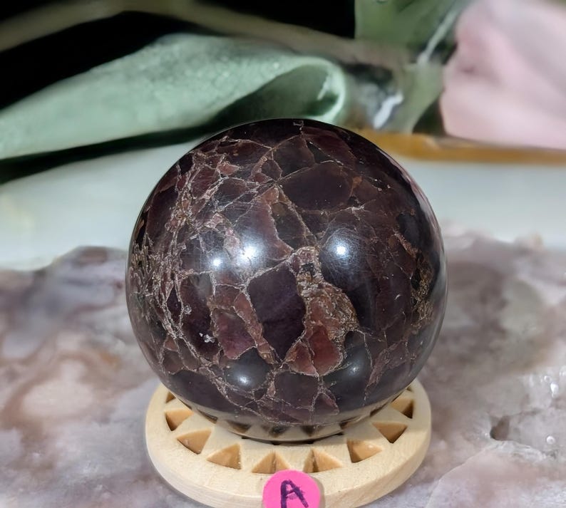 May include: A polished, dark burgundy sphere with a cracked, mosaic-like pattern. The gemstone ball sits on a light wooden stand with triangular cutouts. A small pink sticker with the letter "A" is visible.