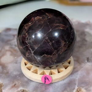 May include: A polished, dark purple sphere with a marbled appearance, resting on a light-colored wooden stand. The sphere has a glossy finish and displays natural variations in color and texture. A pink sticker with the letter "A" is attached to the stand.