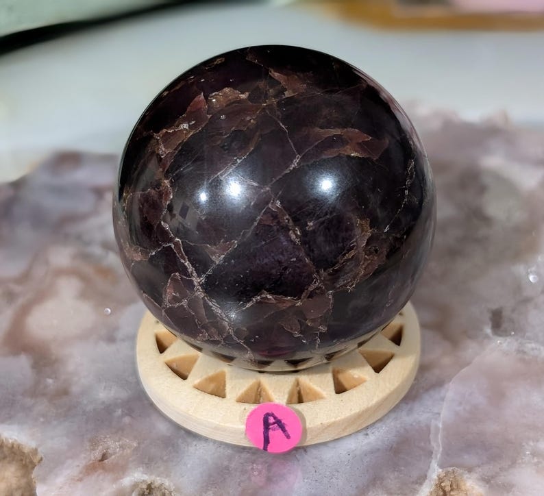 May include: A polished, dark purple sphere with a marbled appearance, resting on a light-colored wooden stand. The sphere has a glossy finish and displays natural variations in color and texture. A pink sticker with the letter "A" is attached to the stand.
