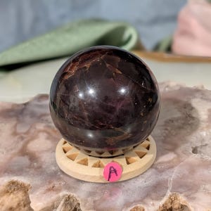 May include: A polished, dark red-brown gemstone sphere. The sphere sits on a small, light-colored wooden stand with a geometric design. A pink sticker with the letter "A" is attached to the stand.