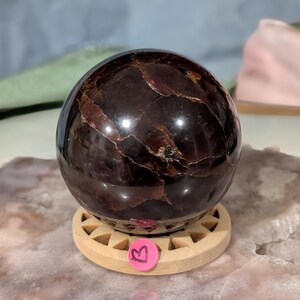 May include: A polished, dark red garnet sphere with visible internal patterns, resting on a small, light-colored wooden stand. The sphere has a glossy finish and is displayed on a light-colored surface. A pink sticker with the letter "B" is on the stand.