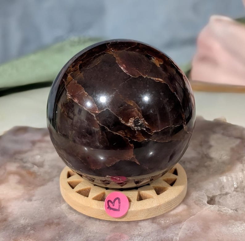 May include: A polished, dark red garnet sphere with visible internal patterns, resting on a small, light-colored wooden stand. The sphere has a glossy finish and is displayed on a light-colored surface. A pink sticker with the letter "B" is on the stand.