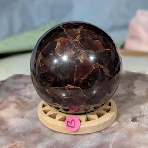 May include: A polished, dark red-brown gemstone sphere with a unique, textured surface. The sphere sits on a light-colored wooden stand with a circular base. The gemstone has a reflective, glossy finish, and is a decorative item.