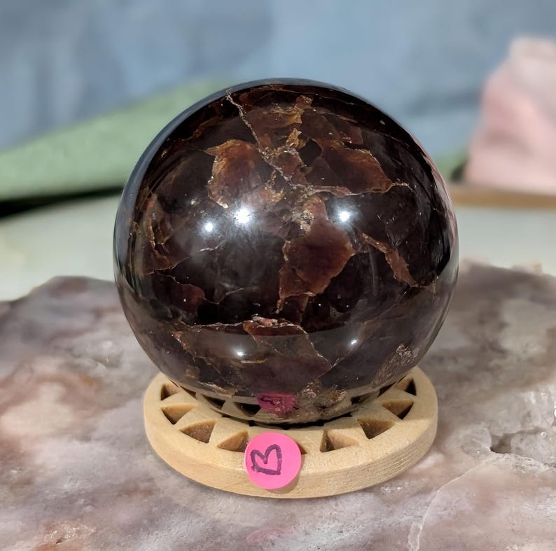 May include: A polished, dark red-brown gemstone sphere with a unique, textured surface. The sphere sits on a light-colored wooden stand with a circular base. The gemstone has a reflective, glossy finish, and is a decorative item.