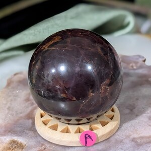 May include: A polished, dark burgundy gemstone sphere, resting on a small, light-colored wooden stand with a geometric design. The sphere has a glossy surface with natural variations in color and texture. A pink sticker with the letter "A" is attached to the stand.