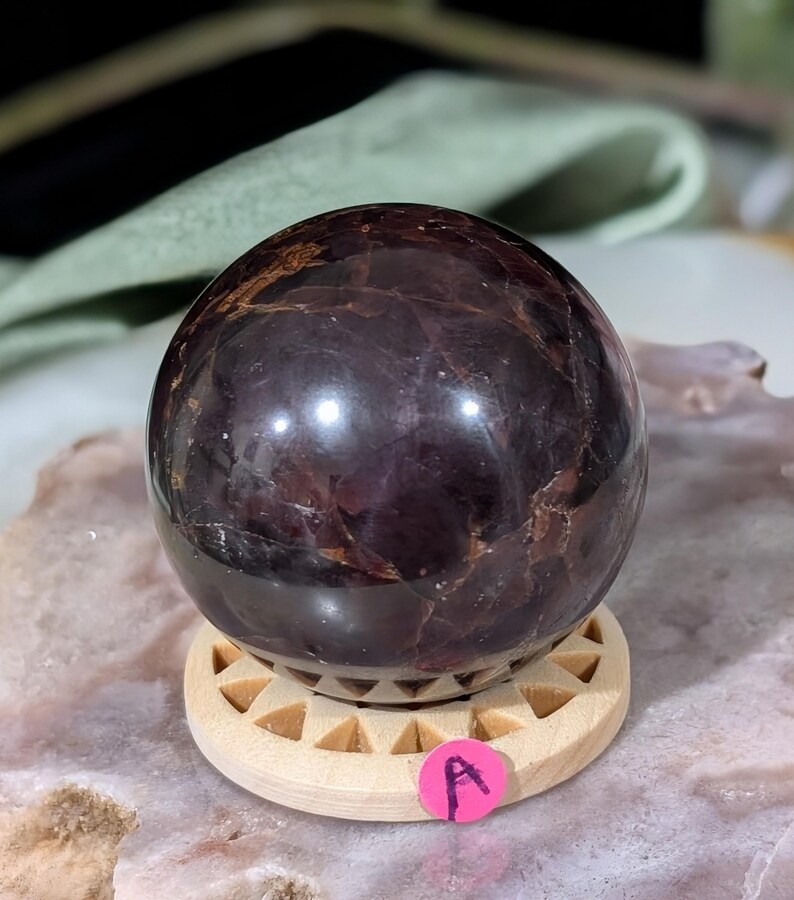 May include: A polished, dark burgundy gemstone sphere, resting on a small, light-colored wooden stand with a geometric design. The sphere has a glossy surface with natural variations in color and texture. A pink sticker with the letter "A" is attached to the stand.