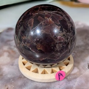 May include: A dark burgundy sphere with a polished surface, resting on a light wooden stand. The sphere has a marbled appearance with lighter veins and inclusions. A small pink sticker with the letter "A" is visible.