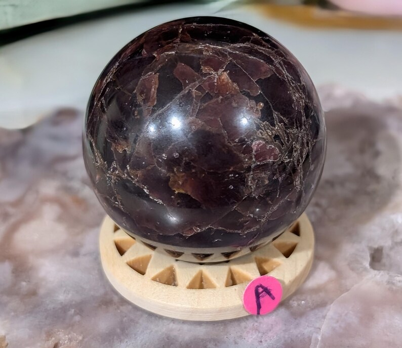 May include: A dark burgundy sphere with a polished surface, resting on a light wooden stand. The sphere has a marbled appearance with lighter veins and inclusions. A small pink sticker with the letter "A" is visible.