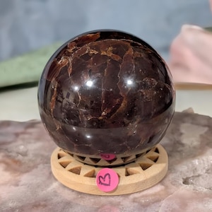 May include: A polished, dark red-brown gemstone sphere with a reflective surface. The sphere sits on a light-colored wooden stand with a circular design. A pink sticker with the letter "B" is attached to the stand.