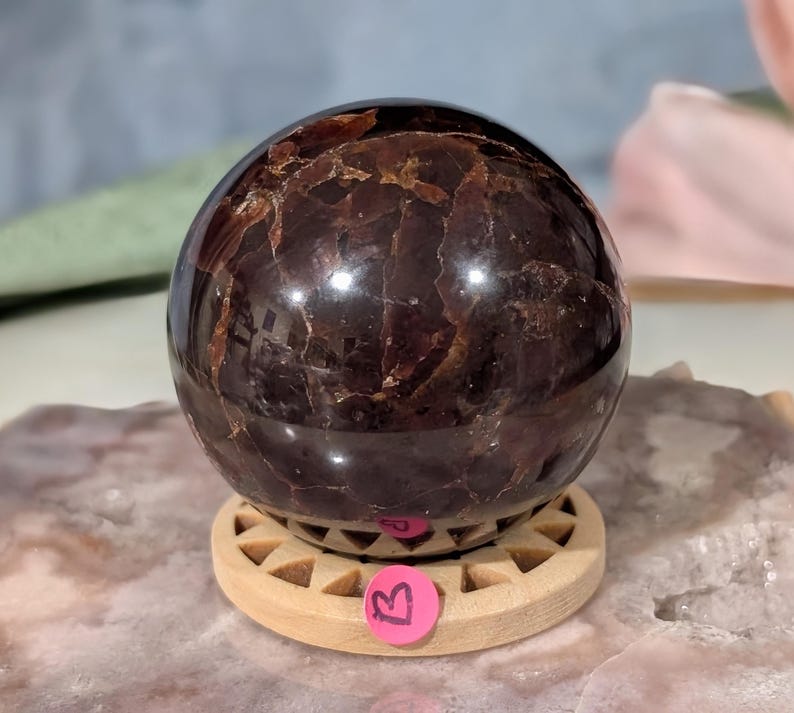 May include: A polished, dark red-brown gemstone sphere with a reflective surface. The sphere sits on a light-colored wooden stand with a circular design. A pink sticker with the letter "B" is attached to the stand.