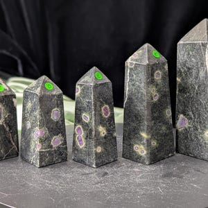 May include: Five dark green obelisk-shaped crystals with purple and light green inclusions. Each crystal has a pointed top with a green sticker. The crystals are arranged in a row on a dark grey surface.
