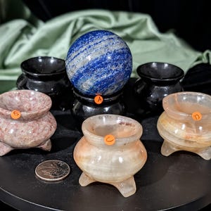 May include: A collection of decorative stone spheres and small, three-legged bowls. A blue and white sphere sits atop a black stand. The bowls are in shades of pink, orange, and white. A coin is included for size reference.