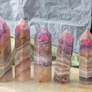 Peut inclure: Cinq tours de cristal d'améthyste avec des étiquettes de prix roses. Les cristaux ont un aspect en couches avec des bandes de violet, de blanc et de marron. Les cristaux sont posés sur une surface de couleur claire avec un fond vert et blanc.