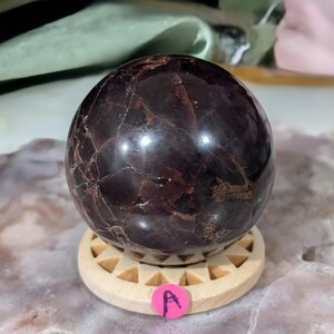 May include: A polished, dark red garnet sphere with natural veining, resting on a light wooden stand. The sphere has a glossy finish and is a deep burgundy color. The stand is round with a geometric design. A pink sticker with the letter "A" is attached to the stand.