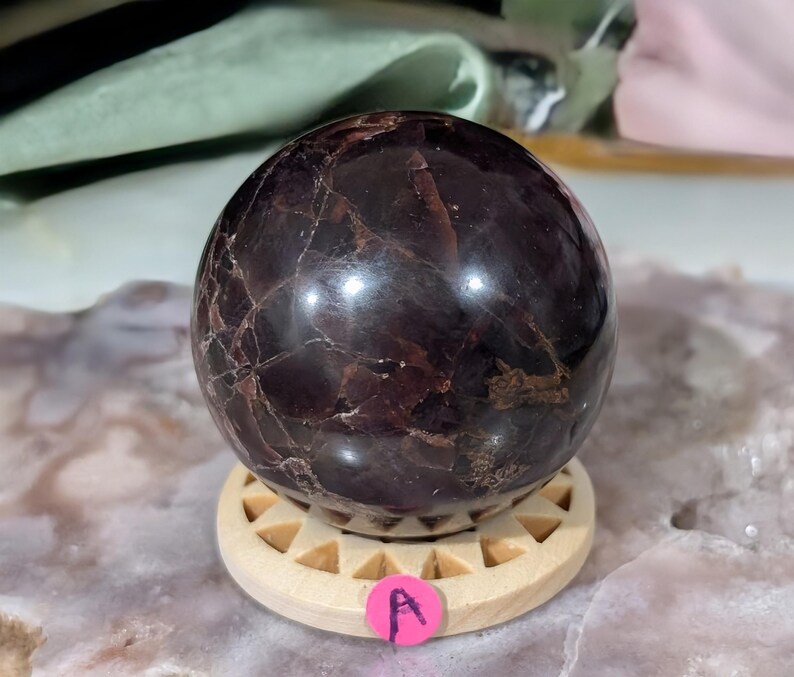 May include: A polished, dark red garnet sphere with natural veining, resting on a light wooden stand. The sphere has a glossy finish and is a deep burgundy color. The stand is round with a geometric design. A pink sticker with the letter "A" is attached to the stand.