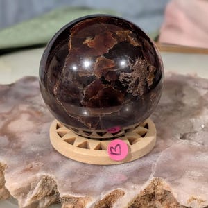 May include: A polished, dark red gemstone sphere with a swirling pattern, resting on a small, light-colored wooden stand. The sphere has a glossy finish and is set on a round stand with triangular cutouts. A pink sticker with the letter "B" is attached to the stand.