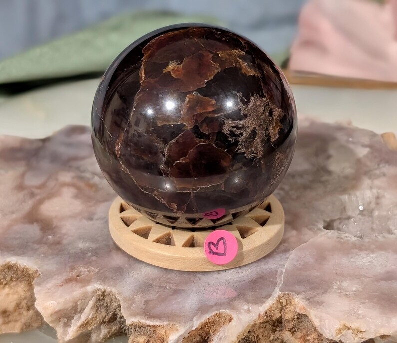 May include: A polished, dark red gemstone sphere with a swirling pattern, resting on a small, light-colored wooden stand. The sphere has a glossy finish and is set on a round stand with triangular cutouts. A pink sticker with the letter "B" is attached to the stand.