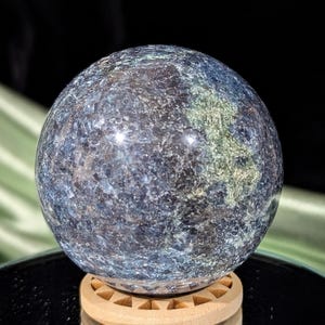 May include: A blue and green sphere made of polished stone, resting on a wooden stand. The sphere has a speckled appearance with a variety of shades of blue and green.