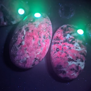 May include: Three pink and black polished stones with a mottled pattern under a UV light. The stones are glowing green.