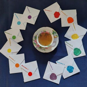 May include: Twelve white envelopes with a different color wax seal on each. The envelopes are arranged in a circle around a cup of tea on a saucer. Each envelope has a date written on it, from December 1st to December 6th.
