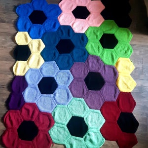 Puede incluir: Una manta de punto colorida hecha de formas hexagonales en varios colores, incluyendo rojo, azul, verde, amarillo y morado. Cada hexágono tiene un círculo negro en el centro.