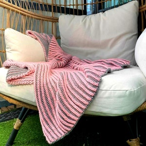 Peut inclure: Un plaid tricoté rayé rose et gris drapé sur une chaise en osier avec un coussin blanc.