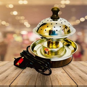 Könnte beinhalten: Ein goldfarbener elektrischer Räucherofen mit einem runden, perforierten Metalldeckel und einem Holzgriff. Der Ofen hat ein schwarzes Kabel und einen Stecker.