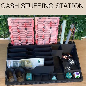 Könnte beinhalten: Ein schwarzer Cash-Stuffing-Organizer mit mehreren Fächern, der rosa und rot gemusterte Budgetblätter, Stifte, Münzen, Würfel und einen Fünf-Euro-Schein enthält. Der Text "CASH STUFFING STATION" befindet sich oben.