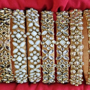 May include: A collection of eight gold-toned bangles, each adorned with intricate floral and geometric patterns. The bangles feature a mix of clear and white embellishments, creating a sparkling effect. The background is a rich, red fabric.