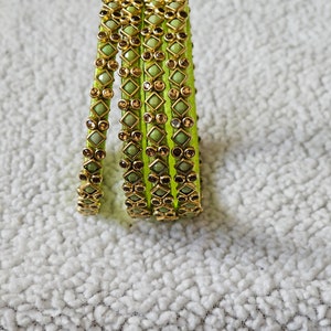 May include: A stack of green bangles with gold-colored accents and decorative gemstones. The bangles are a vibrant green color and feature a repeating pattern of square and round embellishments. The jewelry is displayed on a textured, white surface.