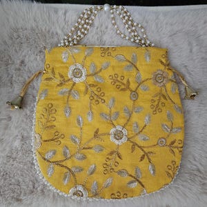 May include: A mustard yellow drawstring pouch featuring intricate silver and white floral embroidery. The bag includes a handle made of multiple strands of white faux pearls and gold-toned beads, with matching gold-toned metal bell-shaped tassels.