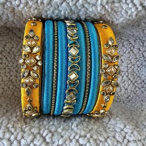 May include: A set of blue and yellow silk thread bangles with decorative embellishments. The bangles are adorned with small, sparkling stones and intricate designs.