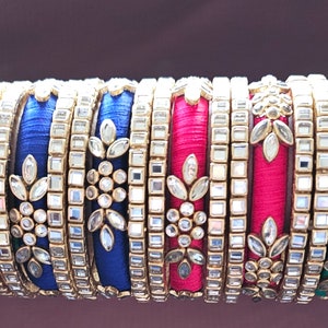 May include: A stack of colorful bangles with intricate designs. The bangles are made of silk thread and decorated with clear rhinestones and gold accents.