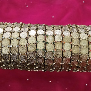 May include: Gold-toned bracelet featuring rows of coin-like embellishments. The bracelet is adorned with small, colorful gemstones. The design is set against a pink background, suggesting a cultural or ethnic jewelry piece.